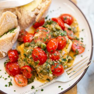cod with tomatoes and capers served on small white plate with bread and silver fork